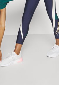 Leggings blu navy con accenti bianchi e verdi, caratterizzati da una striscia laterale e un logo. Scarpe da ginnastica bianche con suole rosa. Design minimalista.