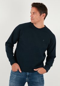 Tengerészkék pulóver puha anyagból, hosszú raglán ujjakkal, kerek nyakkivágással és egy kis logóval a mellrészen. Farmernadrággal viselve.