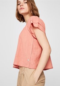 Top corallo chiaro con maniche a sbuffo corte, tessuto testurizzato e orlo arrotondato. Presenta delicate cuciture e un bottone in evidenza.