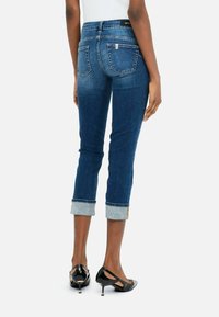 Jean en denim bleu foncé, avec une taille mi-haute, des ourlets roulés avec un revêtement bleu clair, et deux poches arrière. Porté avec des escarpins noirs.