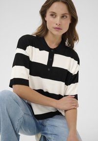 Zwart-wit gestreepte polo met korte mouwen, voorzien van een kraag, een knoopsluiting met drie knopen en ribgebreide manchetten. Geraadpleegd met lichtblauwe jeans.