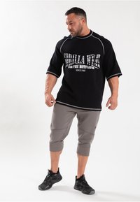 Zwarte shirt met korte mouwen en vetgedrukte witte tekst, gecombineerd met grijze cropped broek en zwarte sportschoenen. Bevat contrasterende stiksels.