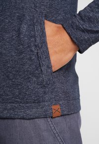 Námořnická modrá, texturovaná pletená mikina s přední kapsou. Má koženou nášivku s logem na lemu. Měkký, ležérní design.