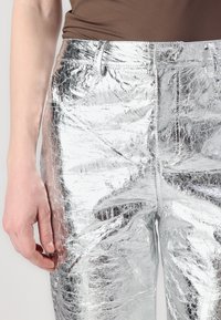 Hopeat metalliset housut, joissa on poimuttunut rakenne, vetoketjukiinnitys, vyölenkit ja tiukka muotoilu, näkyvät vyötäröstä reisiin.