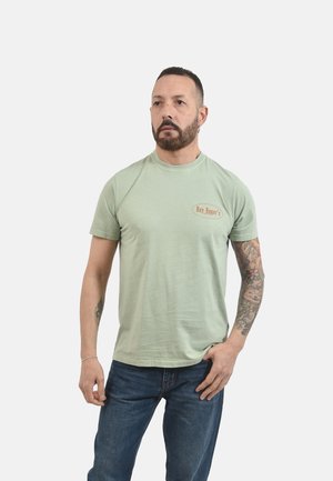 Uomo con barba e tatuaggi che indossa una maglietta verde chiaro "Roy Roger's" e jeans blu, in piedi con una mano in tasca su sfondo bianco.