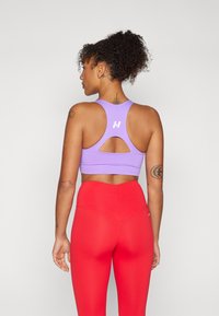 Sujetador deportivo morado con detalle en la espalda y logo, combinado con leggings rojos de cintura alta. Textura suave y diseño ajustado.
