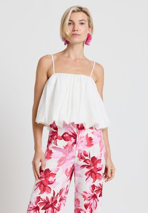 Donna bionda che indossa una canotta bianca ampia senza maniche, pantaloni a gamba larga con fiori rosa e rossi, e grandi orecchini a forma di fiore rosa.