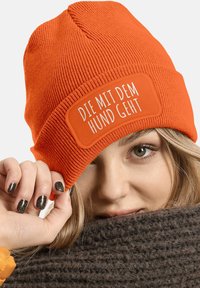 Orange knit beanie with a ribbed texture and a rectangular patch displaying white text. Warm fabric, snug fit, suitable for cold weather.