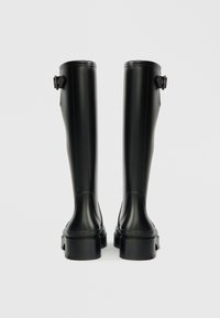 Bottes en caoutchouc noires, hauteur genou, avec une finition lisse, des bouts ronds et un petit détail de boucle près du haut, dotées d'une semelle épaisse.