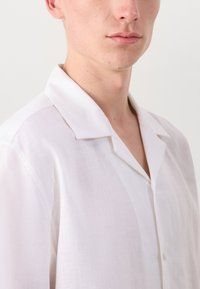 Fehér vászon ing, kivágott gallérral, rövid ujjakkal és egygombos záródással. Az anyag textúrája enyhén áttetsző megjelenésű.