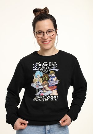 Frau mit Brille, die einen schwarzen Yu-Gi-Oh! GX Sweatshirt trägt, auf dem drei Anime-Charaktere und der Schriftzug "Get Your Game On!" zu sehen sind