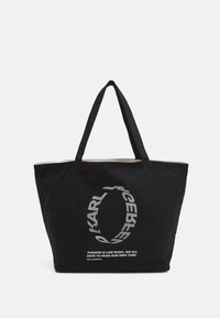 Svart bomullstotepåse med två robusta handtag, prydd med en silverfärgad cirkulär logotyp och text som säger "Mode är som musik..." på framsidan.