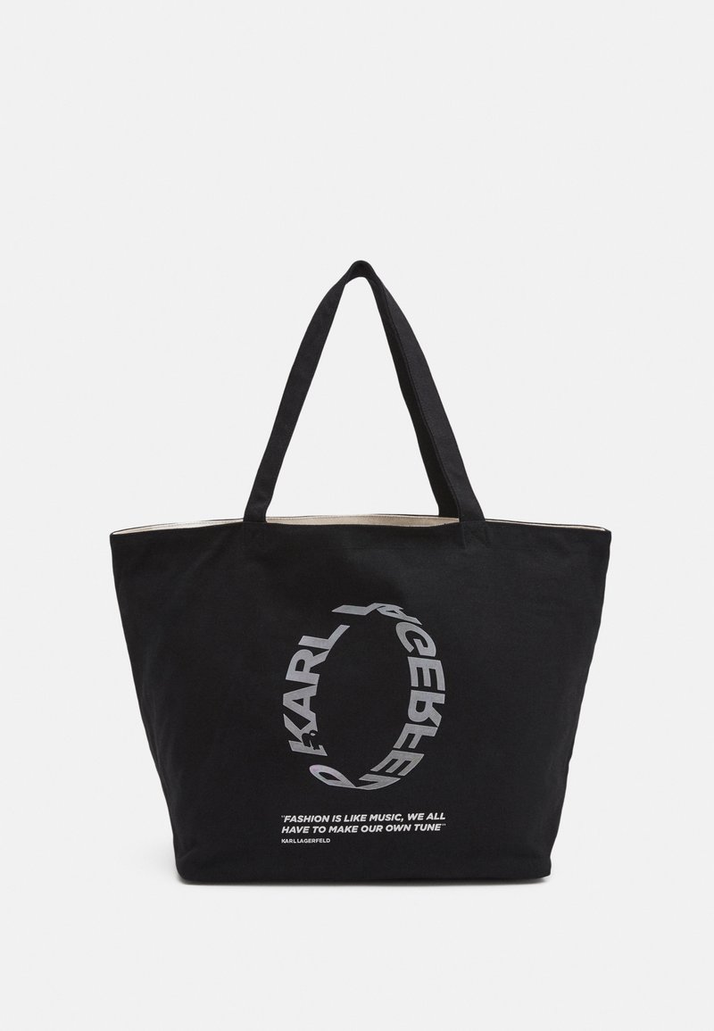 Svart bomullstotepåse med två robusta handtag, prydd med en silverfärgad cirkulär logotyp och text som säger "Mode är som musik..." på framsidan.