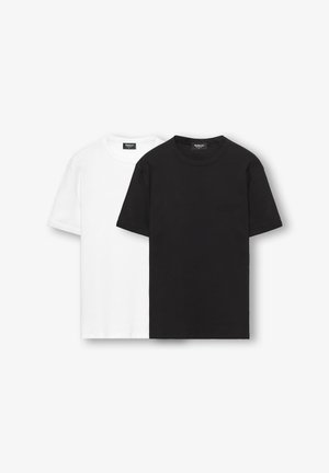 Mies seisoo valkoista seinää vasten, pukeutuneena istuvaan valkoiseen t-paitaan, haalistuneisiin mustiin farkkuihin, mustaan vyöhön ja mustiin loafer-kenkiin, kädet taskuissa.