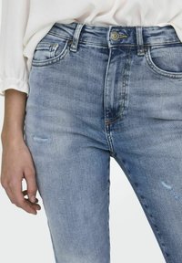 Jeans de mezclilla azul claro con cintura alta, ligera desgaste en el muslo y diseño estándar de cinco bolsillos. Presenta detalles en metal plateado.