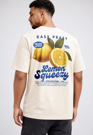 Mann trägt cremefarbenes T-Shirt mit großem Zitronenmotiv und blauem Schriftzug "Easy Peasy Lemon Squeezy" auf dem Rücken, kombiniert mit dunklen Jeans.