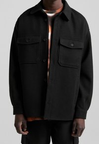 Černá, oversize vlna košile s knoflíkovým zapínáním, dvěma kapsami na hrudi a měkkou texturou. Nositá přes barevné tričko s grafikou.