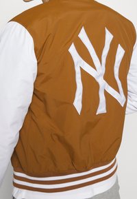 Brun och vit bomberjacka med en stor vit NY-logotyp på baksidan, med randiga ribbade mudd och fåll, slät textur.