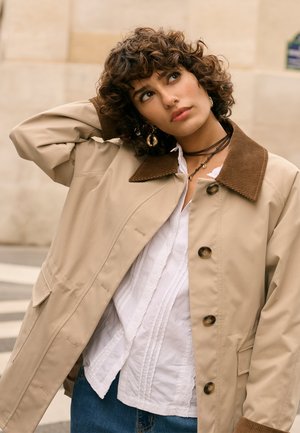 Noor naine lokkis juustega, kellel on seljas beež mantel pruuni kraega, valge särk ja teksad, vaadates mõtlikult ning puudutades oma pead.