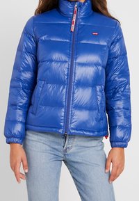 Modrá péřová bunda s vysokým límcem, zapínáním na zip a elastickými manžetami. Disponuje prošívanou texturou a detailem loga na hrudi.