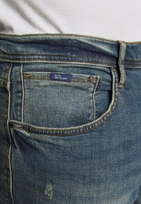 Jeans de mezclilla azul con un aspecto desgastado, que cuentan con detalles de costura, una etiqueta en la cintura y un pequeño bolsillo trasero.