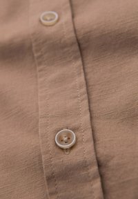 Gros plan d’un tissu de chemise beige avec des coutures visibles et deux boutons beiges translucides fixés le long de la patte de boutonnage.