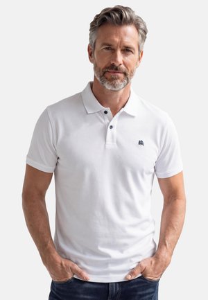 Mittelalter Mann mit grauem Haar und Bart, trägt ein weißes Poloshirt mit kleinem dunklem Logo, Hände in den Taschen, neutraler Hintergrund.