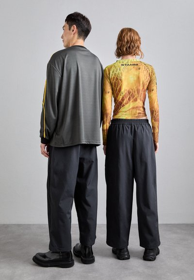 Dwie osoby stoją plecami do siebie. Jedna z nich ma na sobie długą, wzorzystą bluzkę w żółto-brązowe kolory z czarnymi akcentami, a druga szarożółtą w paski. Obie noszą luźne czarne spodnie i wysokie buty.