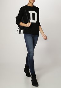 Black sweater with a large grey letter "D," paired with dark blue skinny jeans and black ankle boots. Model carries a fringed black bag.