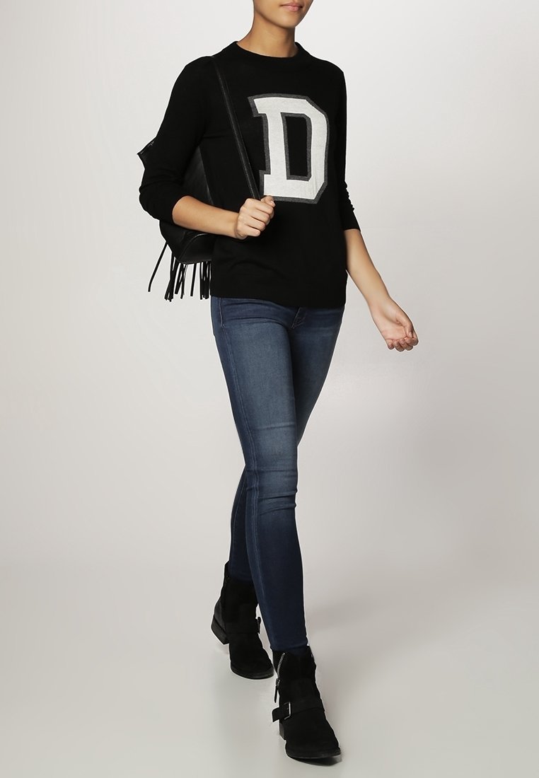 Black sweater with a large grey letter "D," paired with dark blue skinny jeans and black ankle boots. Model carries a fringed black bag.