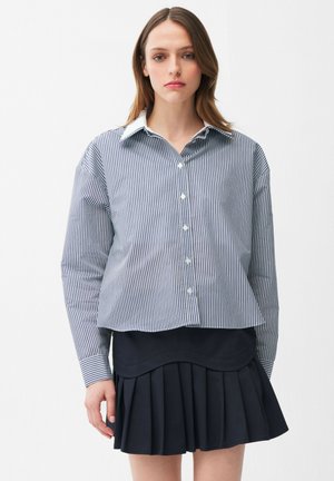 Ung kvinde med glat brunt hår iført en blå og hvidstribet skjorte med knapper og en sort plisseret nederdel mod en hvid baggrund.