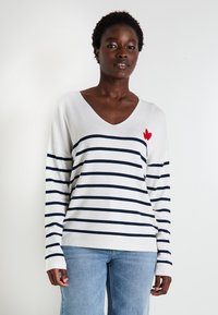 Pull en maille blanc avec des rayures horizontales bleu marine, décolleté en V, et une broderie en forme de cœur rouge sur la poitrine gauche. Manches longues avec des poignets côtelés.