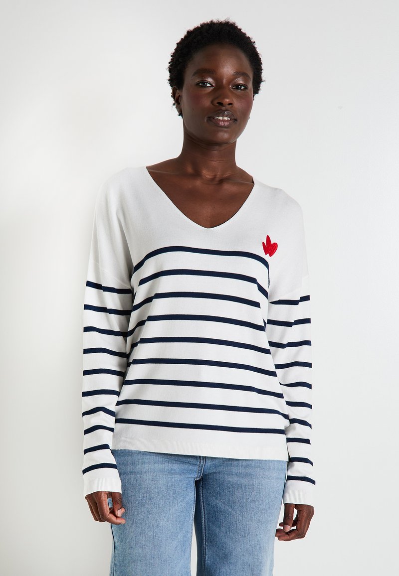 Pull en maille blanc avec des rayures horizontales bleu marine, décolleté en V, et une broderie en forme de cœur rouge sur la poitrine gauche. Manches longues avec des poignets côtelés.