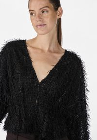 Cardigan nero accorciato con texture frangiata e leggera lucentezza. Scollatura a V e dettaglio di chiusura con bottoni, progettato con una vestibilità ampia.