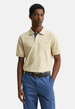 Jonge man die een beige poloshirt draagt met blauwe accenten en een geborduurd logo, blauwe broek en een bruine riem met een geweven patroon.