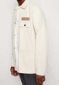 Chaqueta de algodón color crema con cierre de botones, bolsillo en el pecho y parche con el logo. Presenta un corte relajado y un diseño minimalista.