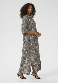 Abito maxi di un leggero beige con motivo a foglie nere, con maniche corte e uno spacco laterale, abbinato a sandali neri con tacco.