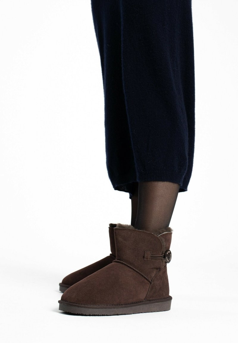 Bottines en daim brun avec un bout arrondi, des détails cousus et un accent de bouton. Assorties à un pantalon noir ajusté et des collants noirs.