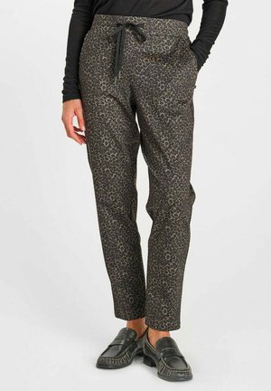 Vrouw die nauwsluitende leopardprintbroeken draagt met een trekkoord in de taille en zwarte instapschoenen, staand met één hand in de zak.