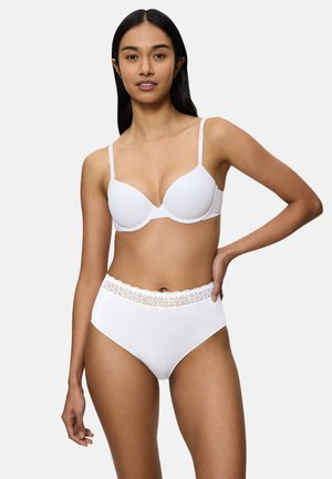 Vrouw draagt een witte beha en hoge slip met kanten rand, staand met één hand op de heup tegen een effen achtergrond.