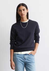 Marineblauwe trui van zachte stof met geribde boorden en zoom, gedragen over een lichte crèmekleurige blouse, gecombineerd met lichtblauwe jeans. Zilveren ketting.