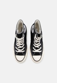 Baskets montantes Converse noires avec lacets blancs, embouts en caoutchouc et écussons de logo visibles sur les parties intérieures des chevilles.