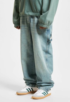 Light blue denim baggy pants with a side pocket, paired with white and gray sneakers featuring green stripes. Textured fabric with visible stitching.
