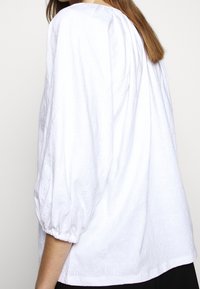 Biała bawełniana bluzka z bufiastymi rękawami, teksturowanym kwiatowym wzorem oraz marszczoną karczkiem z tyłu. Długość poniżej bioder, luźny krój.