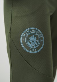 Olīvu zaļas sporta bikses ar teksturētu audumu. Pamanāms zils Manchester City logo augšstilbā ar kuģa un ziedu dizainu.