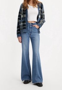 Jean évasé en denim bleu, taille haute, associé à un cardigan à carreaux noir et bleu et un crop-top blanc. Tissu texturé.