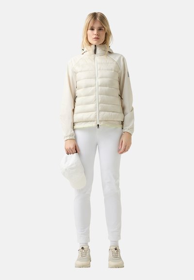 Lysfarvet, puffy jakke med lynlås, lange ærmer og stående krave. Modellen holder en hvid kasket, har hvide bukser på og beige sko.