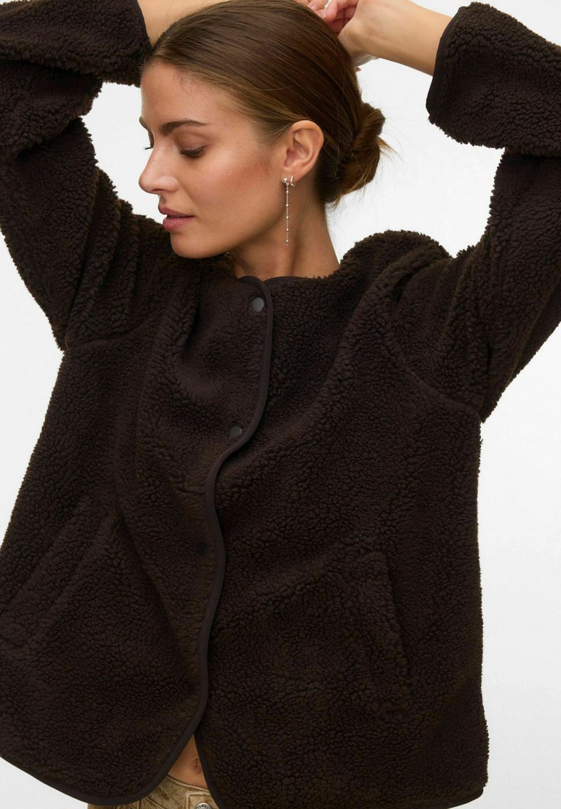 Sieviete, kas pieskārās matiem, valkājot tumši brūnu, strukturētu flīsa jaku ar pogām un garām kuloniem aiz vienkārša fona.