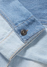 Tejido de mezclilla azul claro con una textura distintiva, que presenta una sección azul oscura y un botón de metal con la marca en el cuello.
