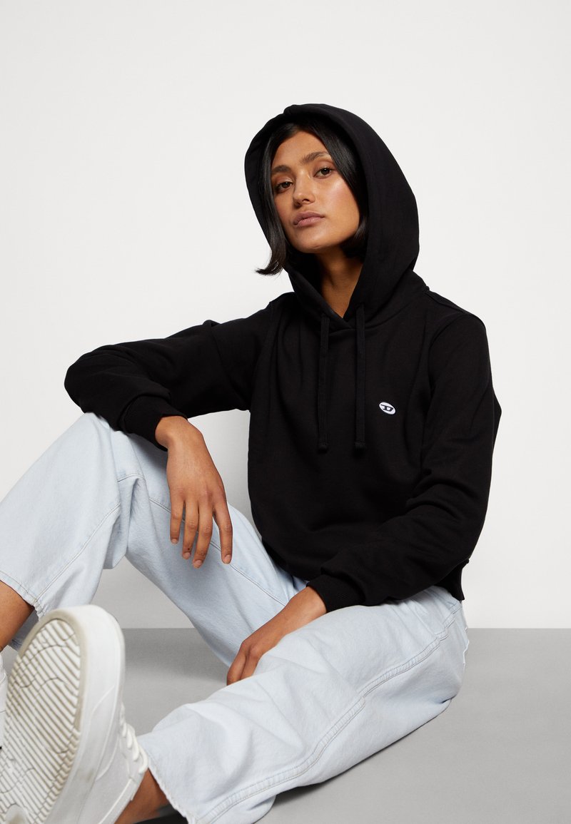 Schwarzer Kapuzenpullover mit Kordelzug und kleinem Logo auf der Brust. Getragen mit hellblauen, weit geschnittenen Jeans und weißen Turnschuhen. Einfaches Design.